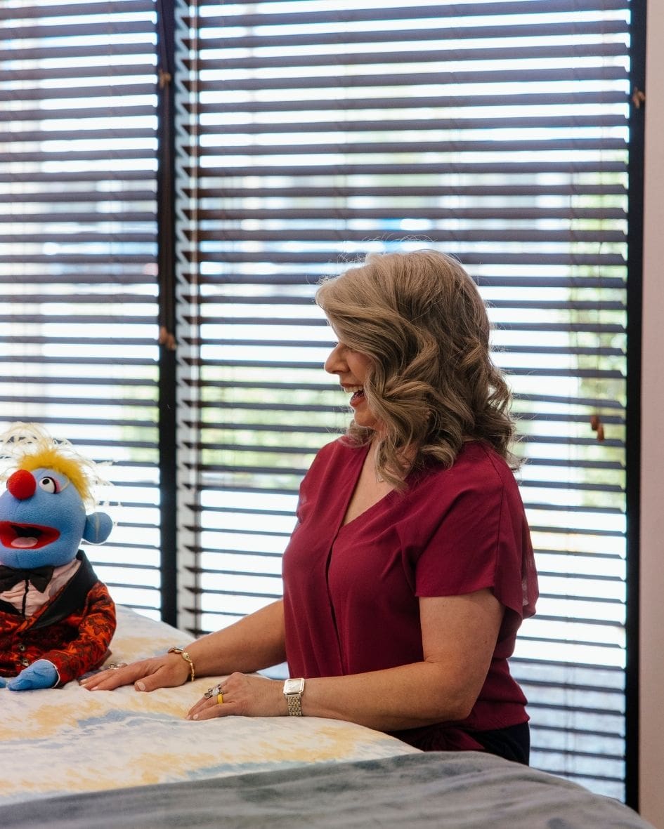 Amy Barilla, pediatric Reiki practitioner, at a table with a colorful puppet she uses with kids